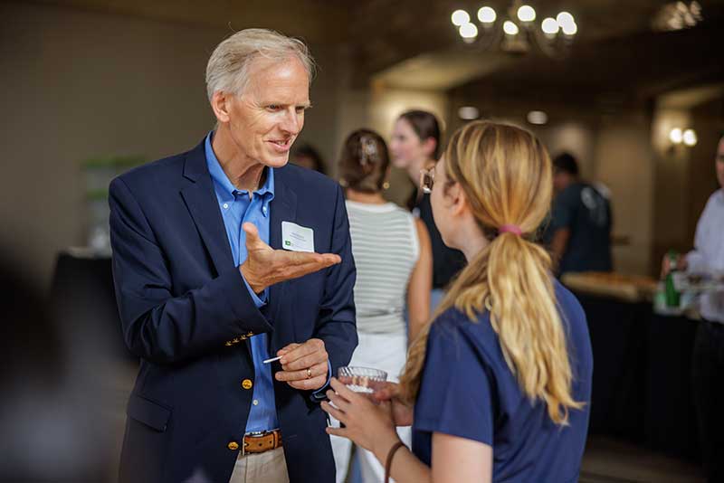 A man and woman has a conversation. The man is making hand gestures as he speaks to the woman who can only be seen from behind.