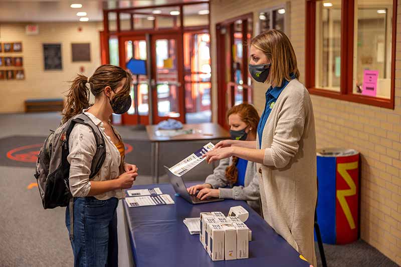 A girl wearing a backpack and mask speaks to  woman, also wearing a mask and pointing to a sheet of paper, about wifi hotspots.