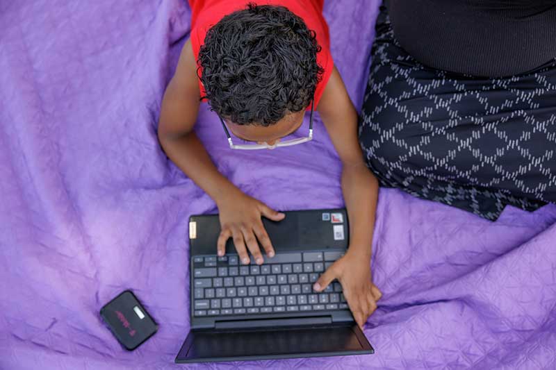 A boy plays on a laptop computer while laying mon a blanket in the grass. A small personal hotspot lays next to the computer.