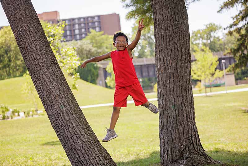 A young boy jumps off of a tree, smiling, and forms a peace sign with his hand.