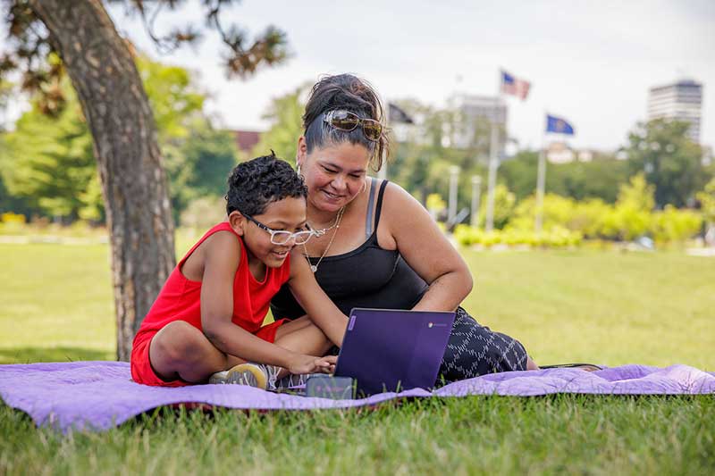 A mother and son sit on a blanket in the grass. The sun wearing glasses plays on a laptop computer while the mother watches.