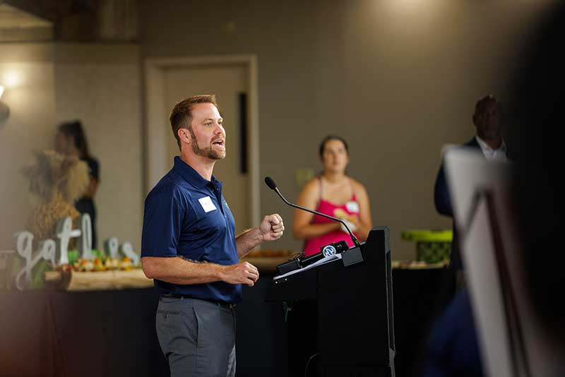 Andrew Wiand, a man wearing a navy polo shirt, speaks at a podium while making fist hand gestures.