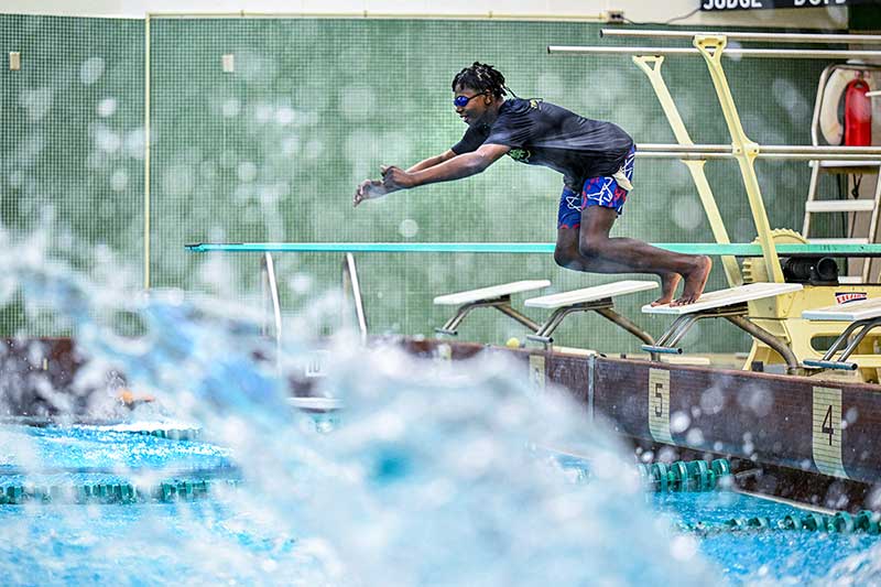 A middle schooler jumps off of a diving board into a pool. In the foreground is a out of focus splash.