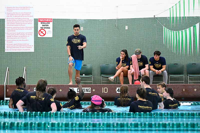 Middle schoolers stand in a pool and listen to a student athlete who is outside of the pool. Three other athletes sit in chairs.