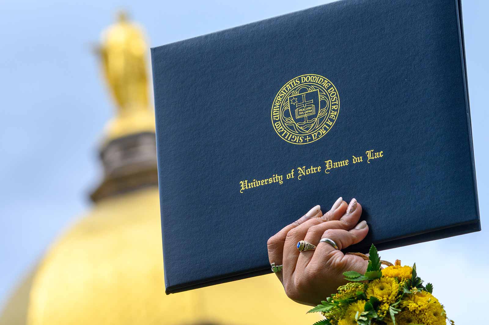 In the background the Golden Dome is out of focus. In the foreground a close up of someone's hand, wearing rings and a wrist corsage, holds onto a blue leather Univerty of Notre Dame diploma folder.