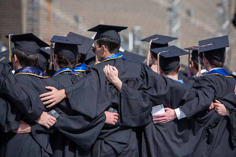 Gradutes wearing caps and gowns stand arm in arm with their backs to the camera.