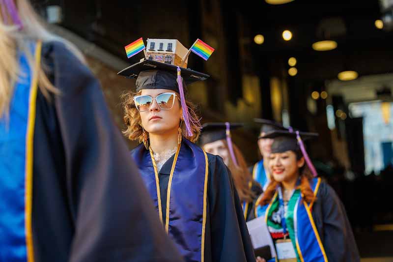 A woman walks out from Notre Dame's Stadium tunnel wearing a graduation cap decorated with pride flags and a building.