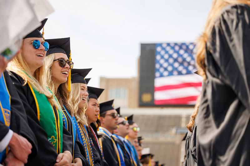 Graduates wearing caps and gowns standing looking right. In the background a stadium videoboard displays an american flag.