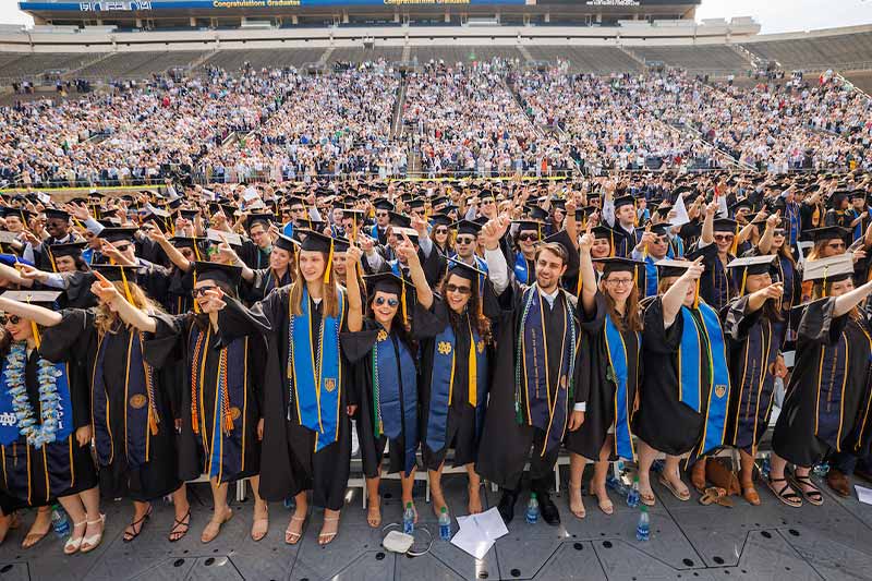 Graduates stand in the stadium, with thousands of fans behind them in the stands, singing together raising their arms in the air.