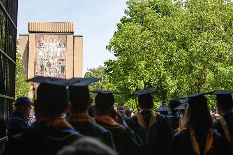 Graduates wearing caps and gowns in the foreground, blurred, facing Touchdown Jesus in the background, in focus.