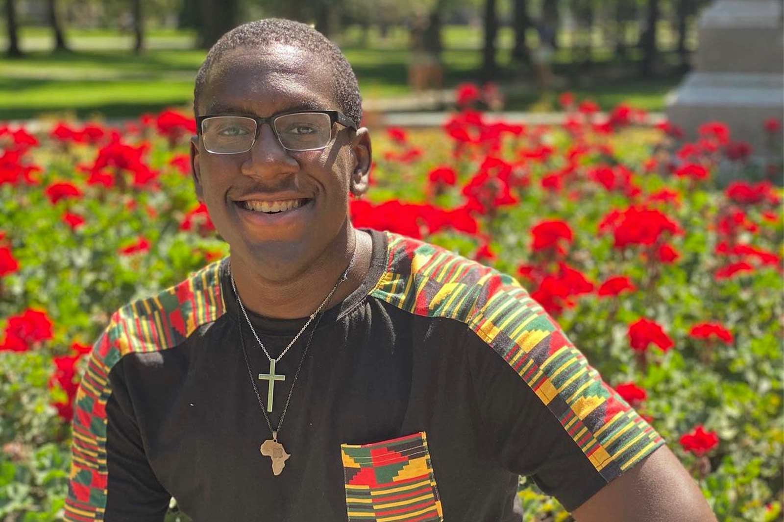 A black student wearing glasses and a two necklaces - one a cross, the other Africa - sits in front of red geraniums at the base of the Jesus statue in front of Main Building on Notre Dame's campus.