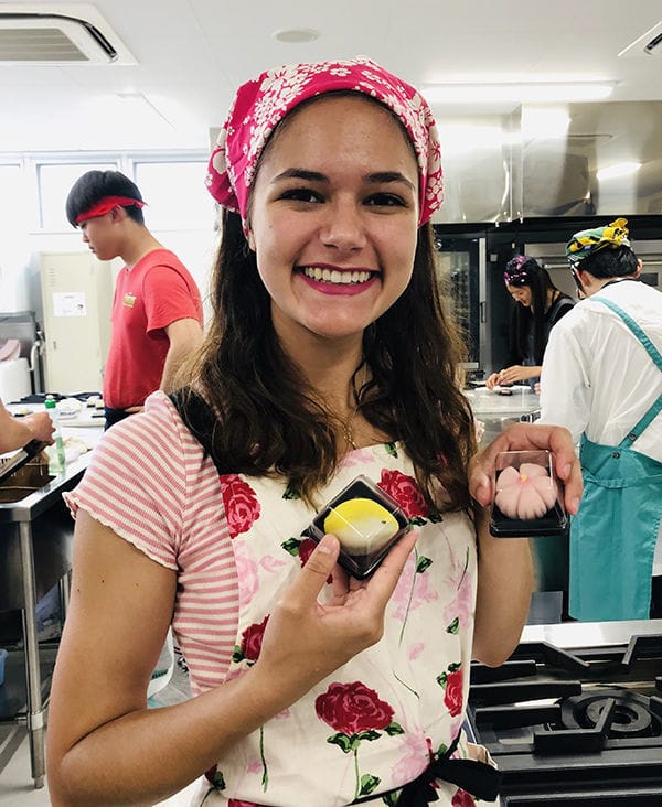 A light skinned girl with her hair pulled back in a bandana holds small pink and yellow baked goods in a kitchen. She is wearing an apron with red and pink roses on it.