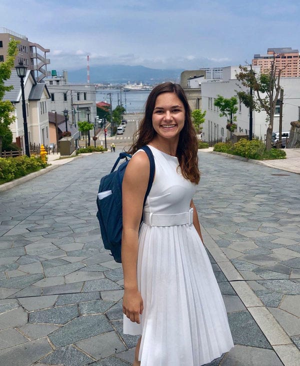 A light skinned girl with dark hair wearing a white dress smiles on a gray paver road with water in the distance.