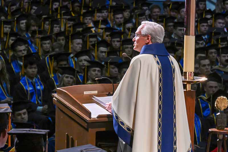 Fr. Jenkins, a White man wearing a white priest robe with a dark blue trimming, speaks in front of a audience in front of a podium during a Mass.