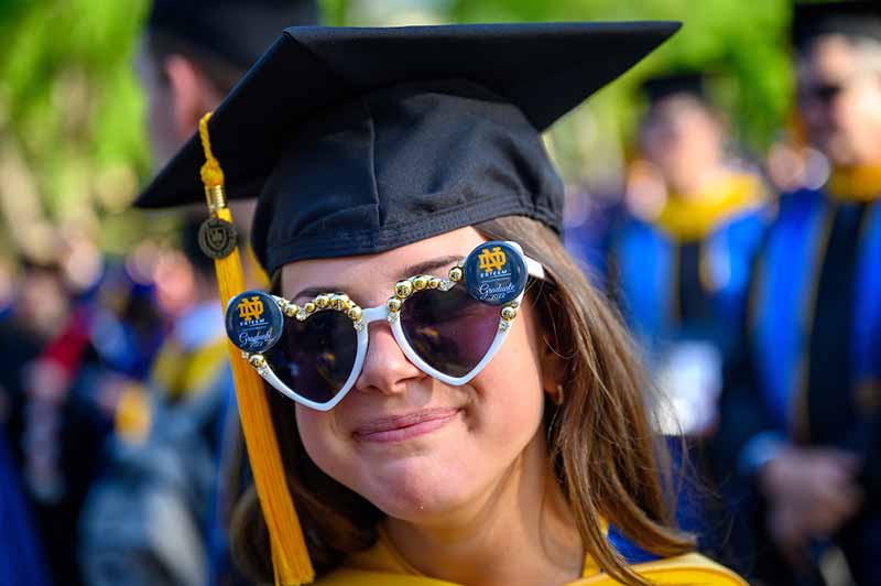 A White female grauate wearing a black cap with a yellow tassel smiles for the camera while wearing large, white heart-shaped sunglasses with Notre Dame buttons on each lens.
