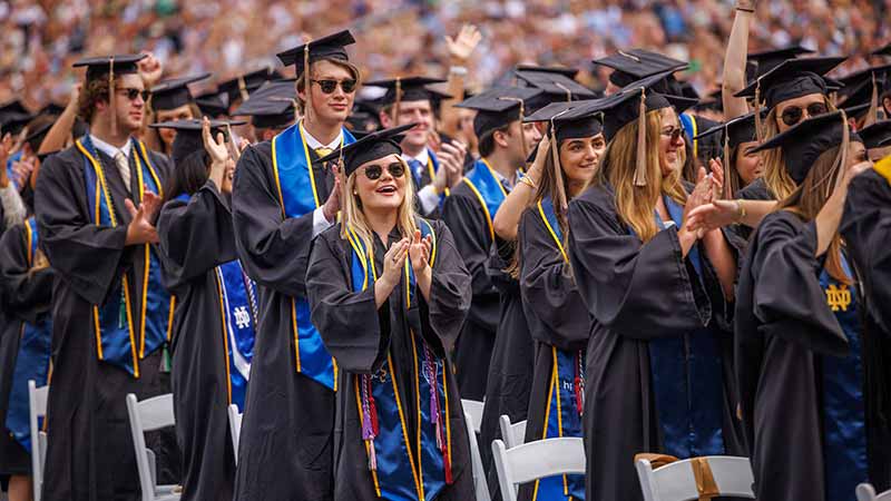 Standing graduates wearing black graduation robes and caps clap and cheer.