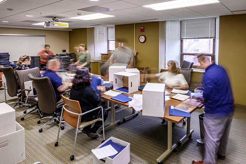 A group of people, some blurred in motion, inside of a conference room stand and sit around a table and prepare diplomas.