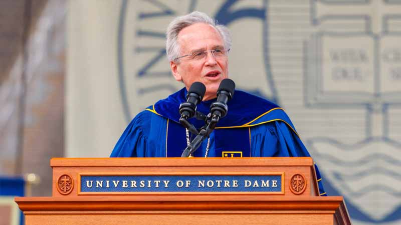 Fr. Jenkins, a White man wearing wearing the navy blue commencement regalia of the University of Notre Dame, speaks at a podium with the seal of the University on it.