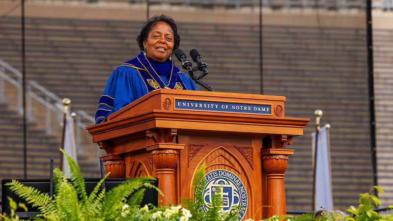 Sharon Lavigne, a Black woman wearing the navy blue commencement regalia of the University of Notre Dame, speaks at a podium with the seal of the University on it.