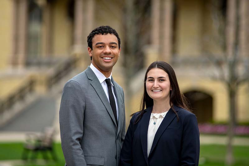 Devin Diggs, a black man, and Morgan La Sala, a white woman, poses for a photo. Devin is wearing a gray suit with a black tie, and Morgan wears a blue blazer and white top.