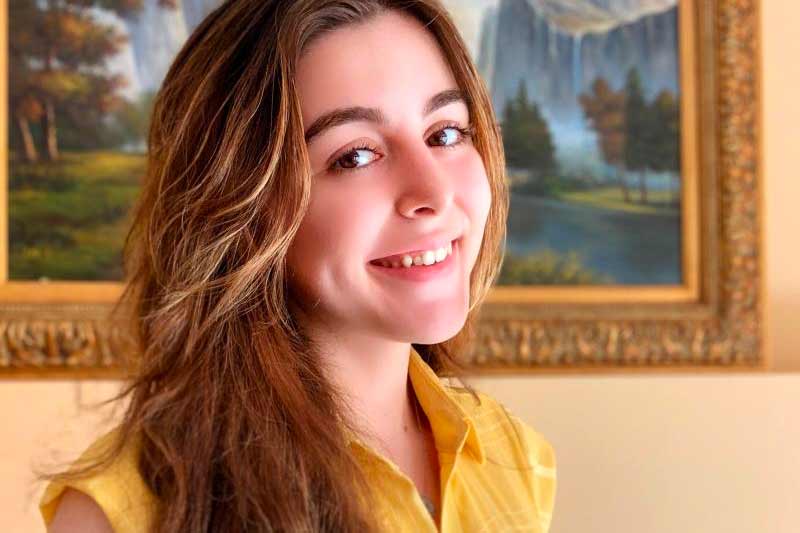 A female student poses for a portrait photo standing in front of artwork hung on a wall behind her.