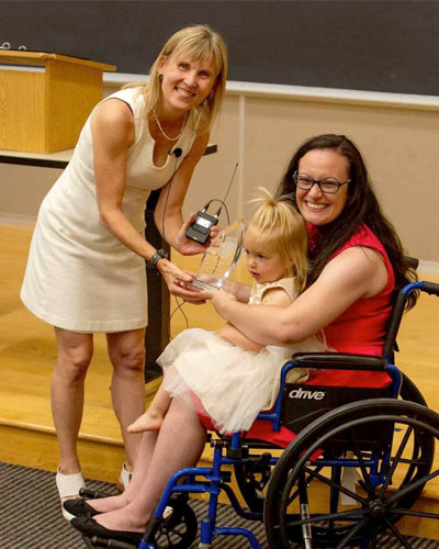 Abby, on the right, sits in her wheelchair with her daughter on her lap. Laura Carlson, on the left, is bending over giving Abby an award.
