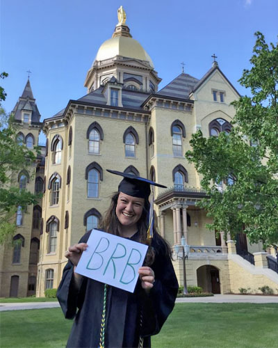 Abby stands in front of the golden home holding a sign that says BRB (Be Right Back).