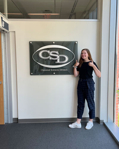 Audrey Miles stands in front of a wall with a sign on it that reads Chemical Sciences Division