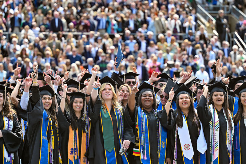 Graduates standing together in the crowd and holding their pointer finger in the air.