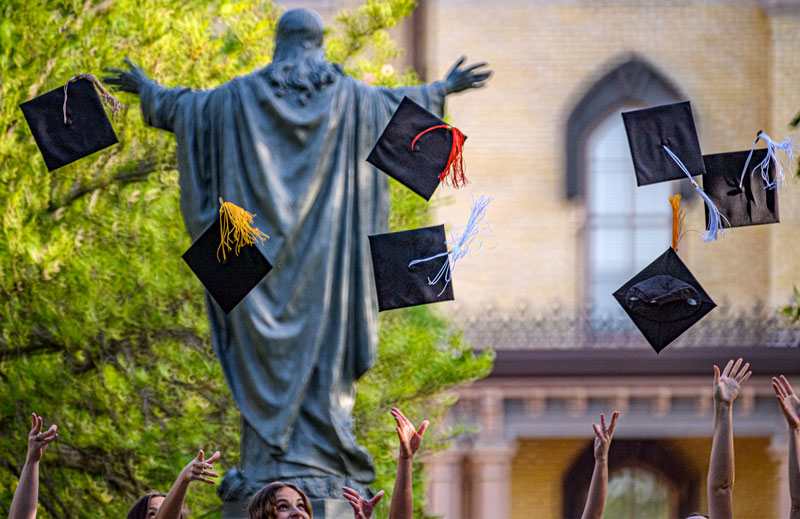 Motion blurred commencement caps fly in the air near the statue of Jesus on the Main quad.