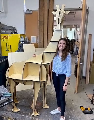 Gabriela stands next to a laser cut giraffe bookshelf.