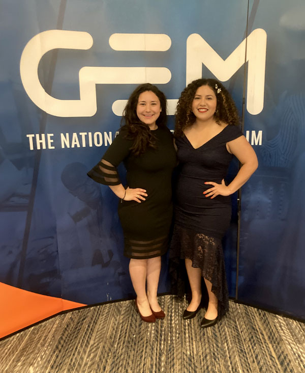 Karen Angeles and a friend stand in front of a sign with the letters G E and M on it.