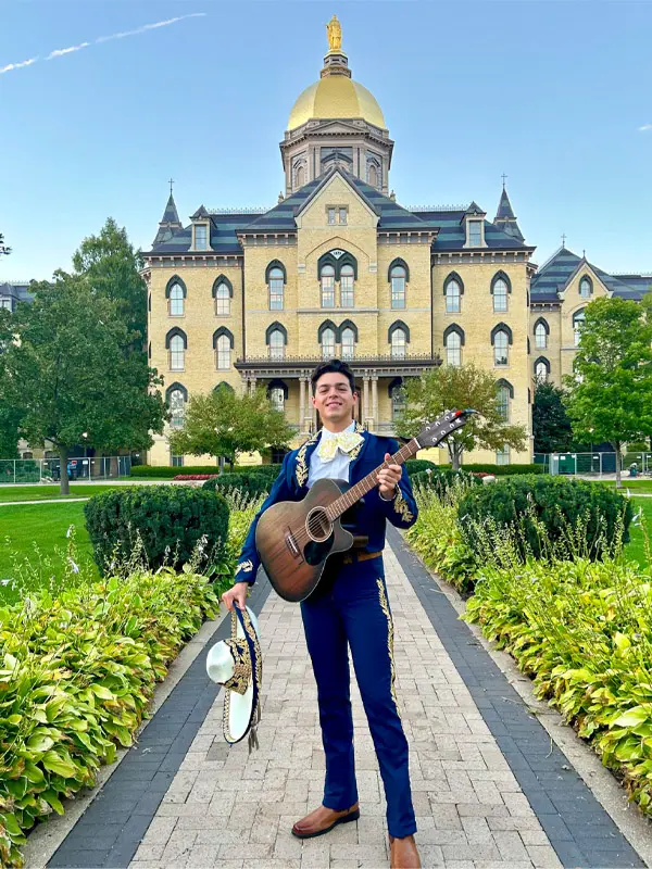 Alan Avalos se para frente a la cúpula vistiendo su traje de mariachi y sosteniendo una guitarra y un sombraro.