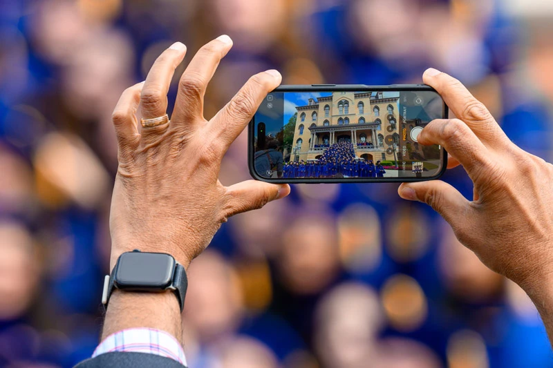 Hands hold a smartphone taking a photo of graduates in blue gowns gathered in front of Morrissey Hall at the University of Notre Dame.  A blurred crowd wearing blue is visible in the background.