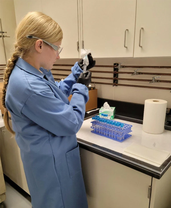 A white female with long blond hair wearing a blue lab coat, safety glasses, and black gloves prepares a syringe in a laboratory setting.  A rack of small, blue-capped test tubes sits on the counter beside them.