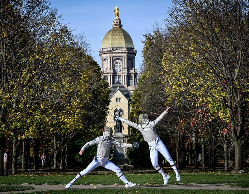 Two fencers in white uniforms and protective masks lunge at each other on a grassy area in front of the Main Building at the University of Notre Dame. The Golden Dome is prominent in the background, and autumn trees frame the scene.