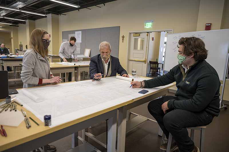 Two students, one standing and pointing at a large piece of paper, discuss with their professor who sits on a stool, all wearing masks.