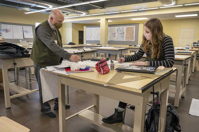 A masked professor talks with a masked female student sitting at a desk inside of a classroom.