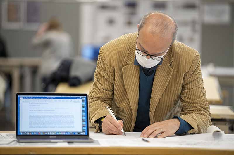 A man wearing glasses, a mask, and a tan corduroy suit jacket leans over a desk and sketches. There is an on and open computer on the desk aside him.