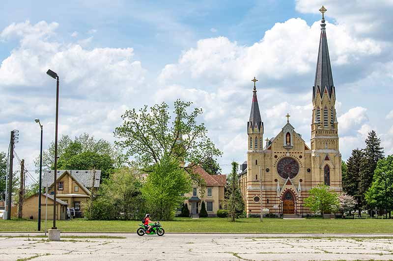 Grass growing out of cracks in an empty parking lot. A single motocyclist drives on the road along two homes and a church with two large steeples.