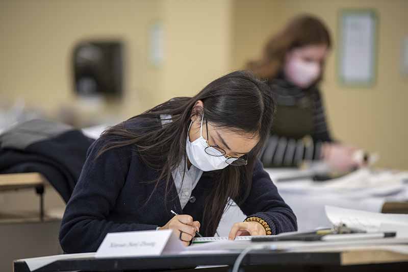 A masked student sketches at a desk in a classroom.