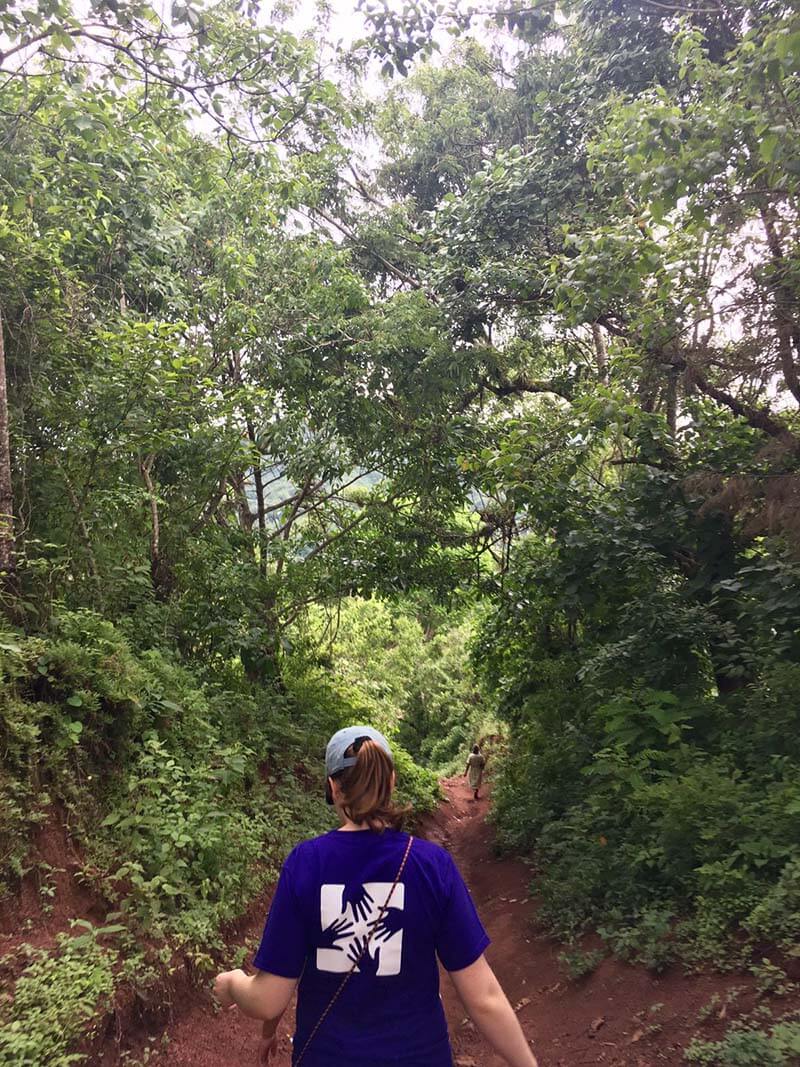 A woman walks down a narrow red dirt trail, surrounded by green trees.