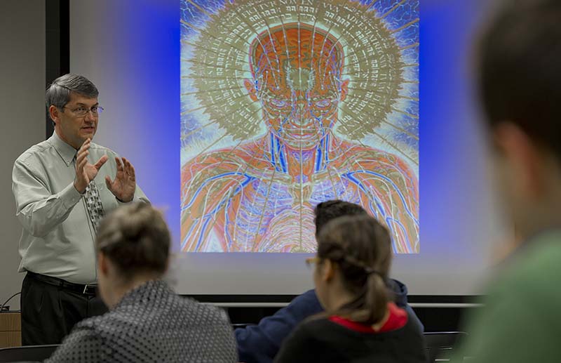 Dom teaches a glass, with an image of a human head and body on the screen behind him.