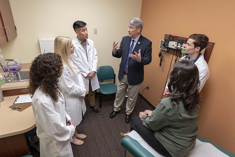 Dom stands in a medical room, with students in lab coats.