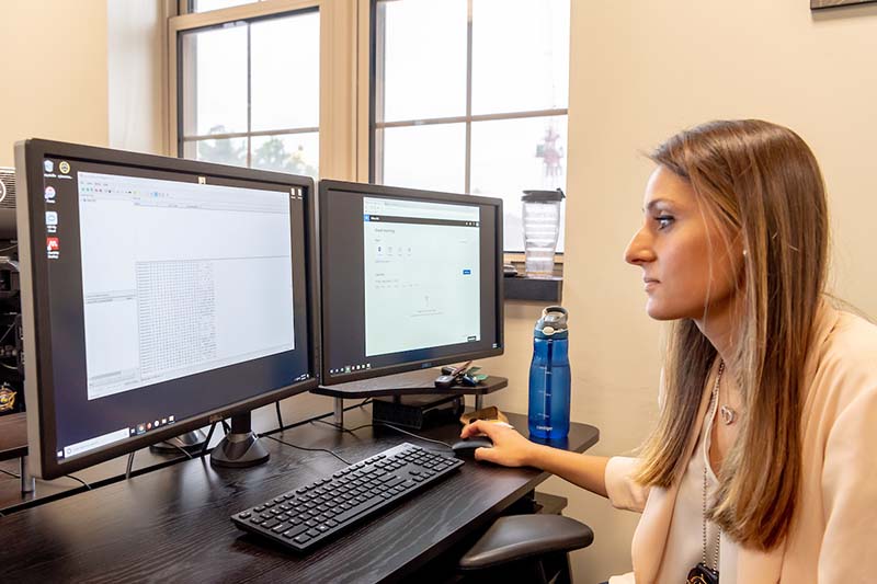 A female student wearing a badge sits at a desk looking at a computer screen.