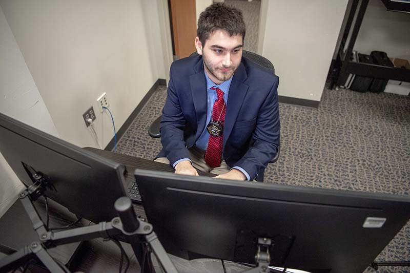 A male student wearing a badge sits at a desk looking at a computer screen.