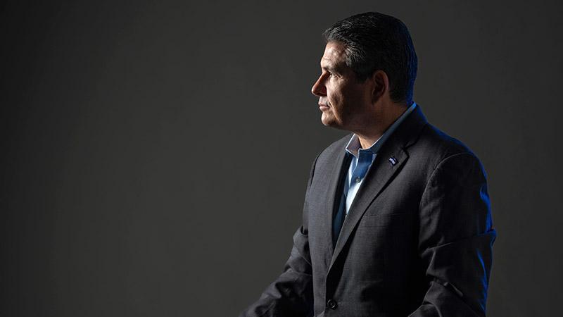 A side portrait of Juan Sebastian Chamorro. He wears a dark suit and is backlit by a blue light set against a grey backdrop.