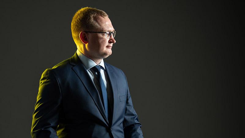 Side portrait of Nazarii Stetsyk. He wears glasses and a dark suit. He is set against a grey backdrop and backlit with a soft yellow light.