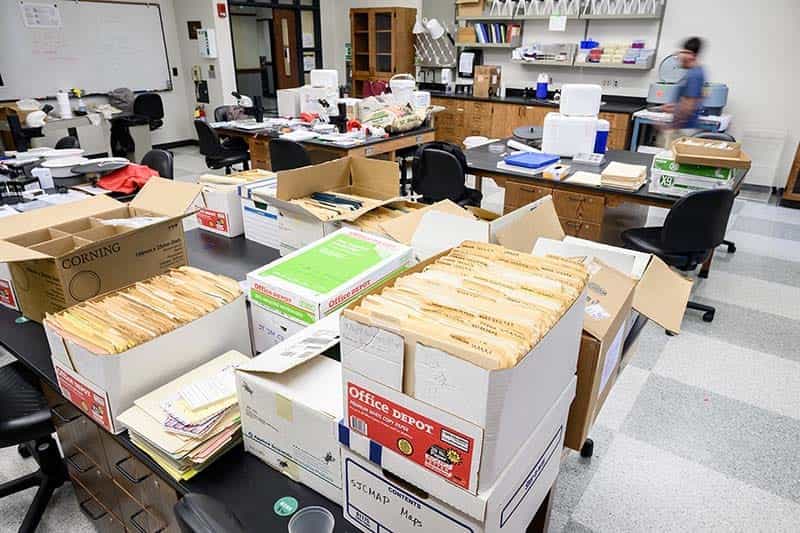 Boxes of manilla folders cover tables in the lab. Microscopes and mosquito trap supplies are on the other tables.