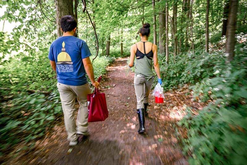 Two people walk a wooded trail carrying mosquito trap supplies.